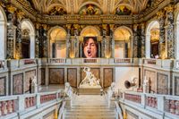 The exuberant staircase of the Kunsthistorisches Museum Vienna.  A large portrait of a screaming woman is mounted between to center columns above the grand stairway. The picture is part of the exhibition communication for Beethoven Moves.