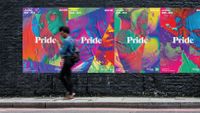 A POC-woman walking on a side way in front of a series of posters advertising Pride Toronto. The posters are a colorful and vibrant collage of different faces with the word Pride in the center.