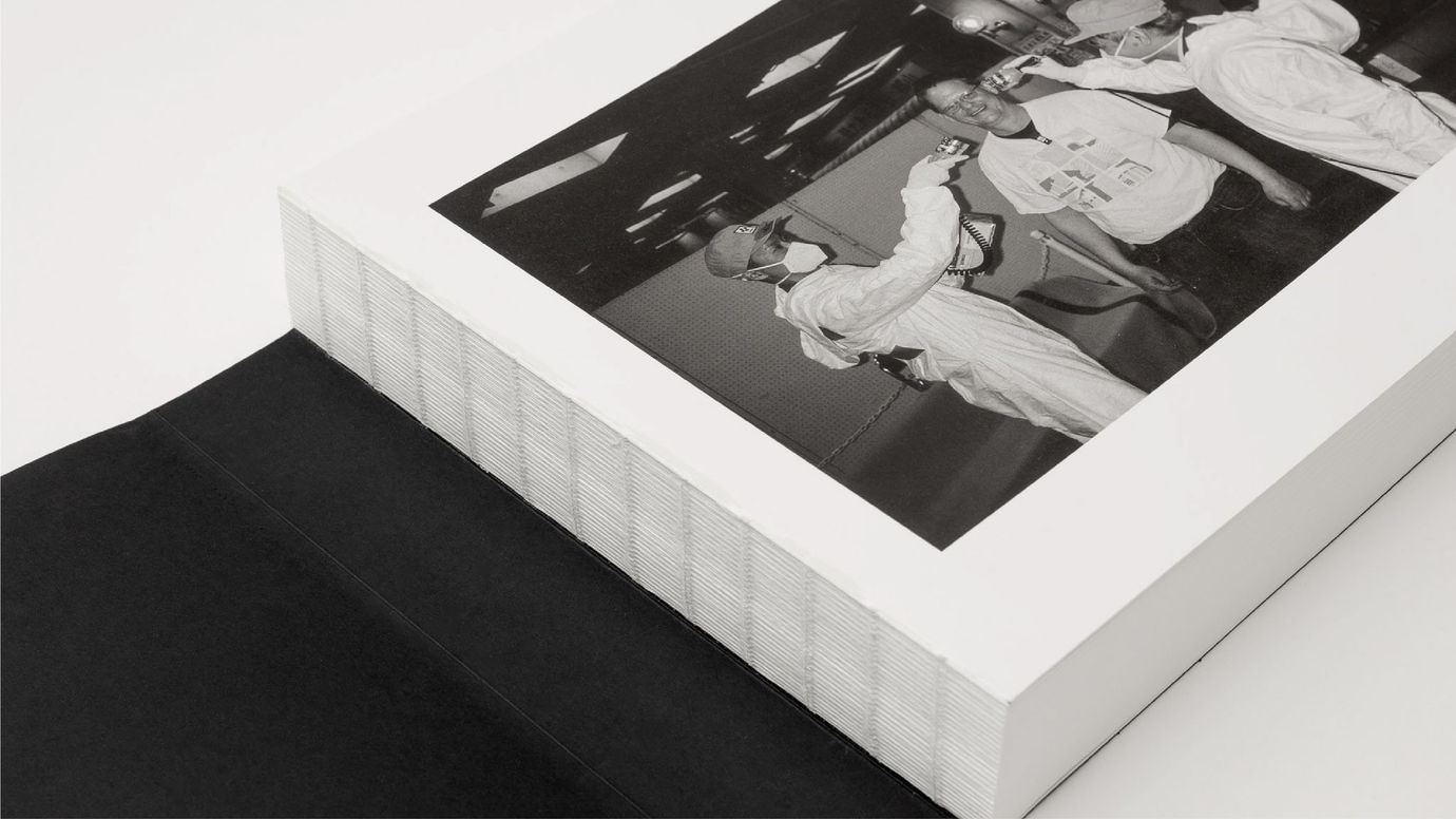 An open black book reveals a view of its thick open spine binding. On the first page, part of an image is visible: a black and white photo depicting a smiling man being scanned with handheld devices by two other men.
